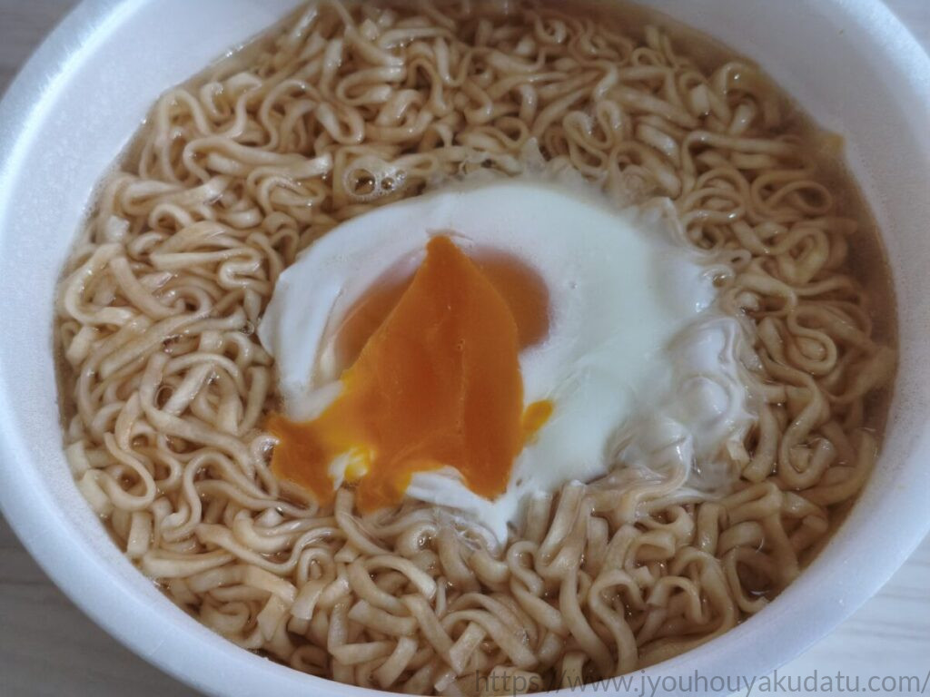 チキンラーメンどんぶりの半熟状の黄身を崩した様子。オレンジ色の黄身が白身と麺の上に広がっている